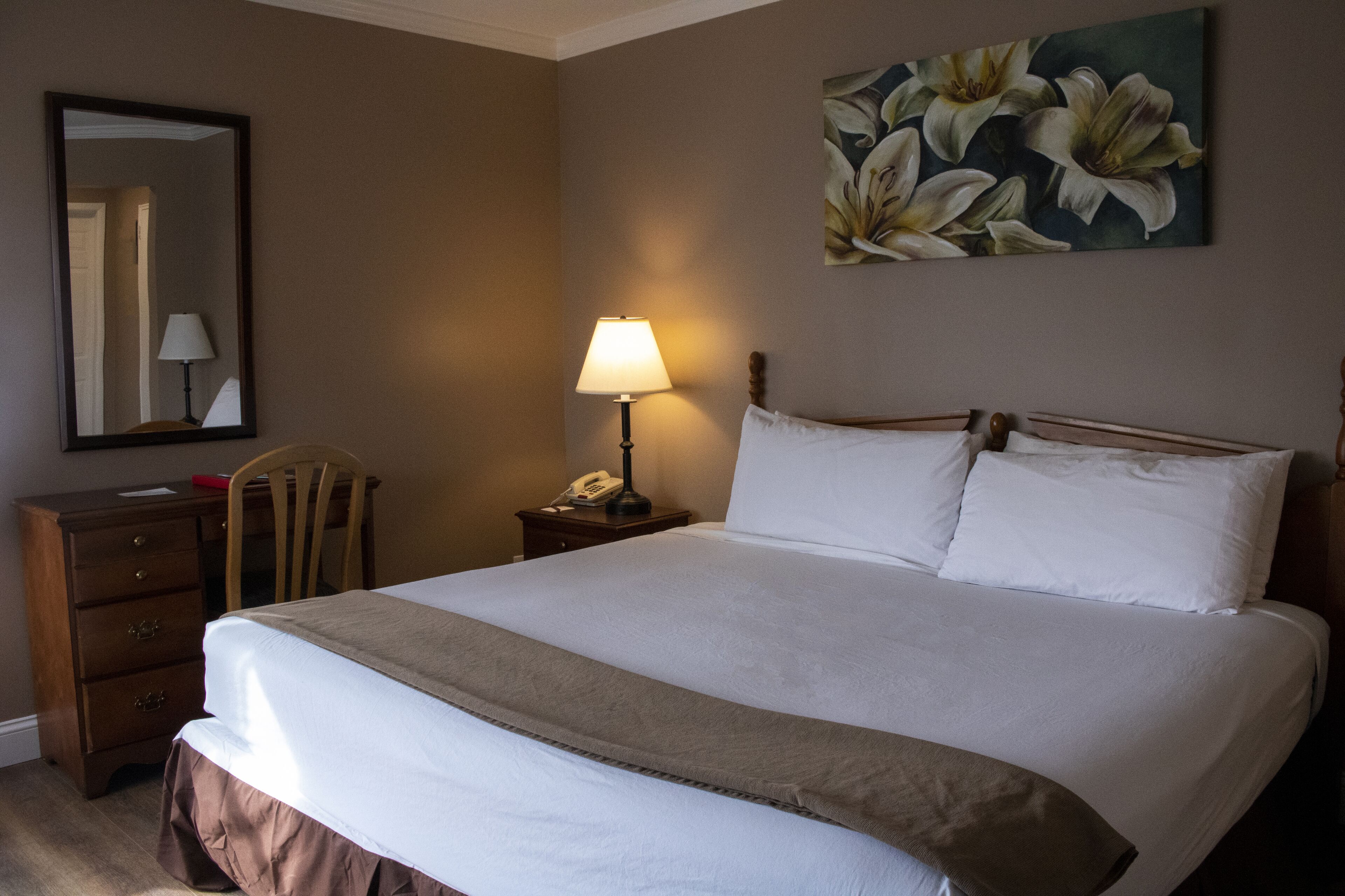 Una cama bien tendida con sábanas blancas, una mesita de noche de madera con una lámpara, una silla de madera y un cuadro de flores en la pared.