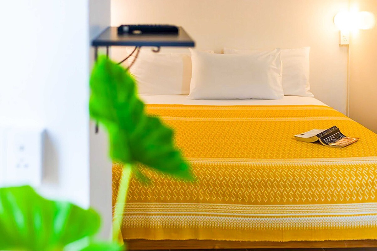A hotel room with a bed, white pillows, a yellow bedspread, a bedside table with a phone, and a wall-mounted light.