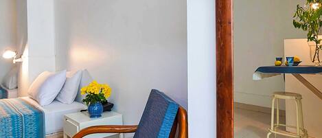 Una habitación con una cama, una silla, una mesita con un florero y un taburete.