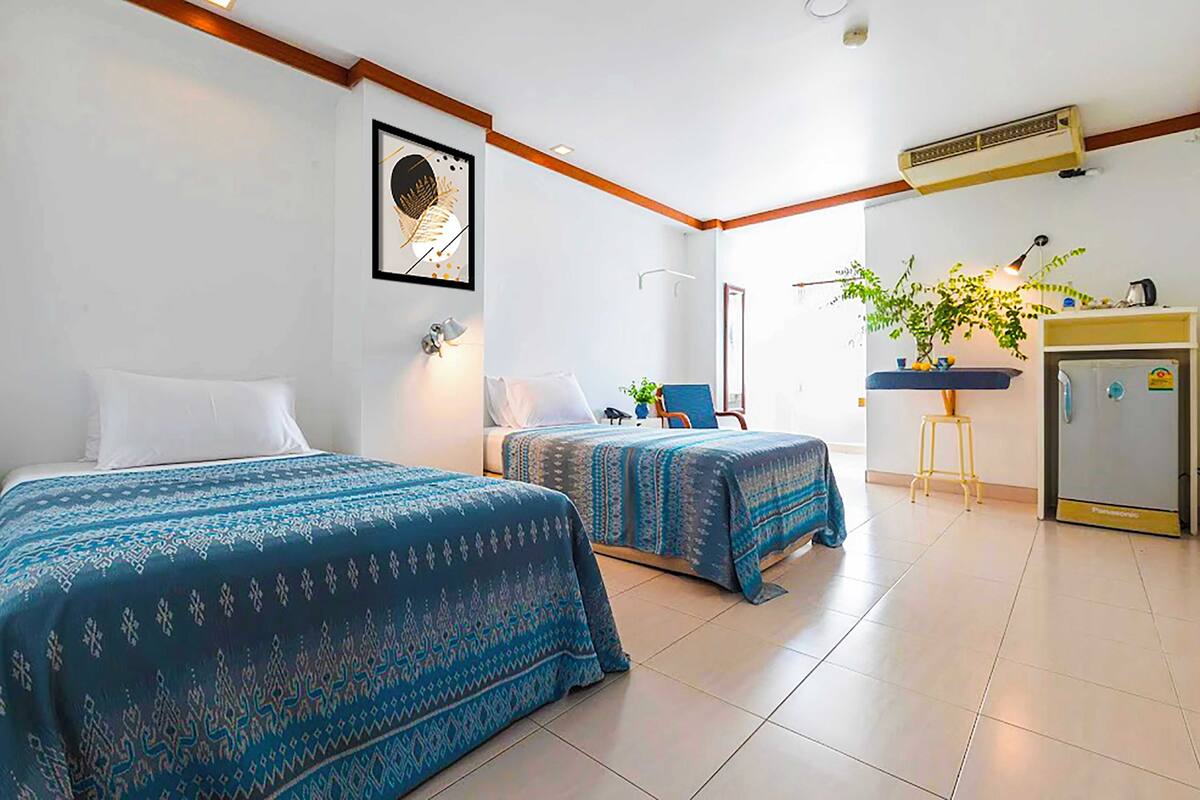 A hotel room with two beds, a desk, a chair, a plant, and a refrigerator.