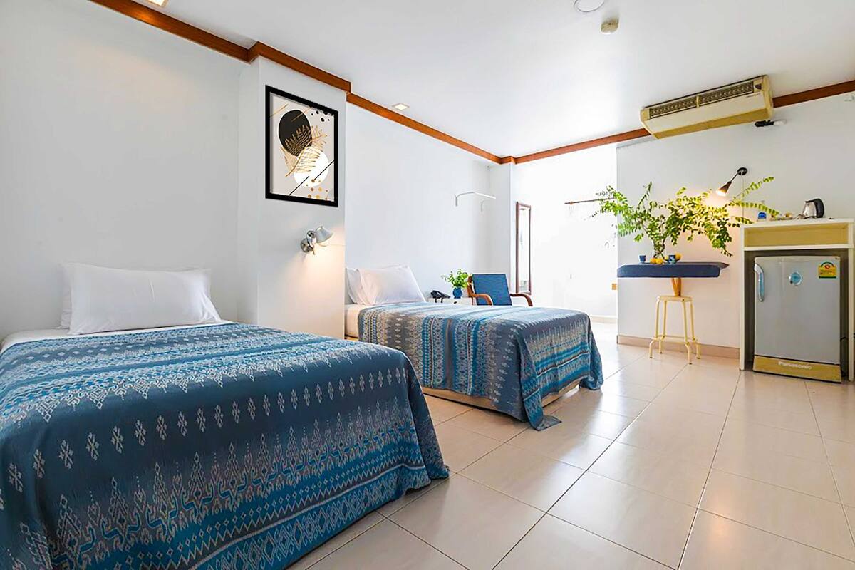 A hotel room with two beds, a desk, a chair, a plant, and a refrigerator.