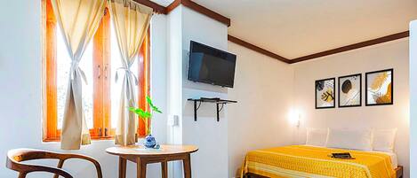 Habitación de hotel con cama, televisión, una mesita, una silla y ventana con cortinas.