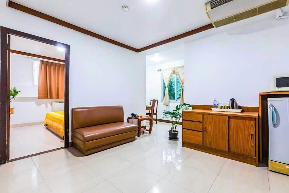 A hotel room with a sofa, a dining table, a chair, a potted plant, a wooden cabinet, and a refrigerator.