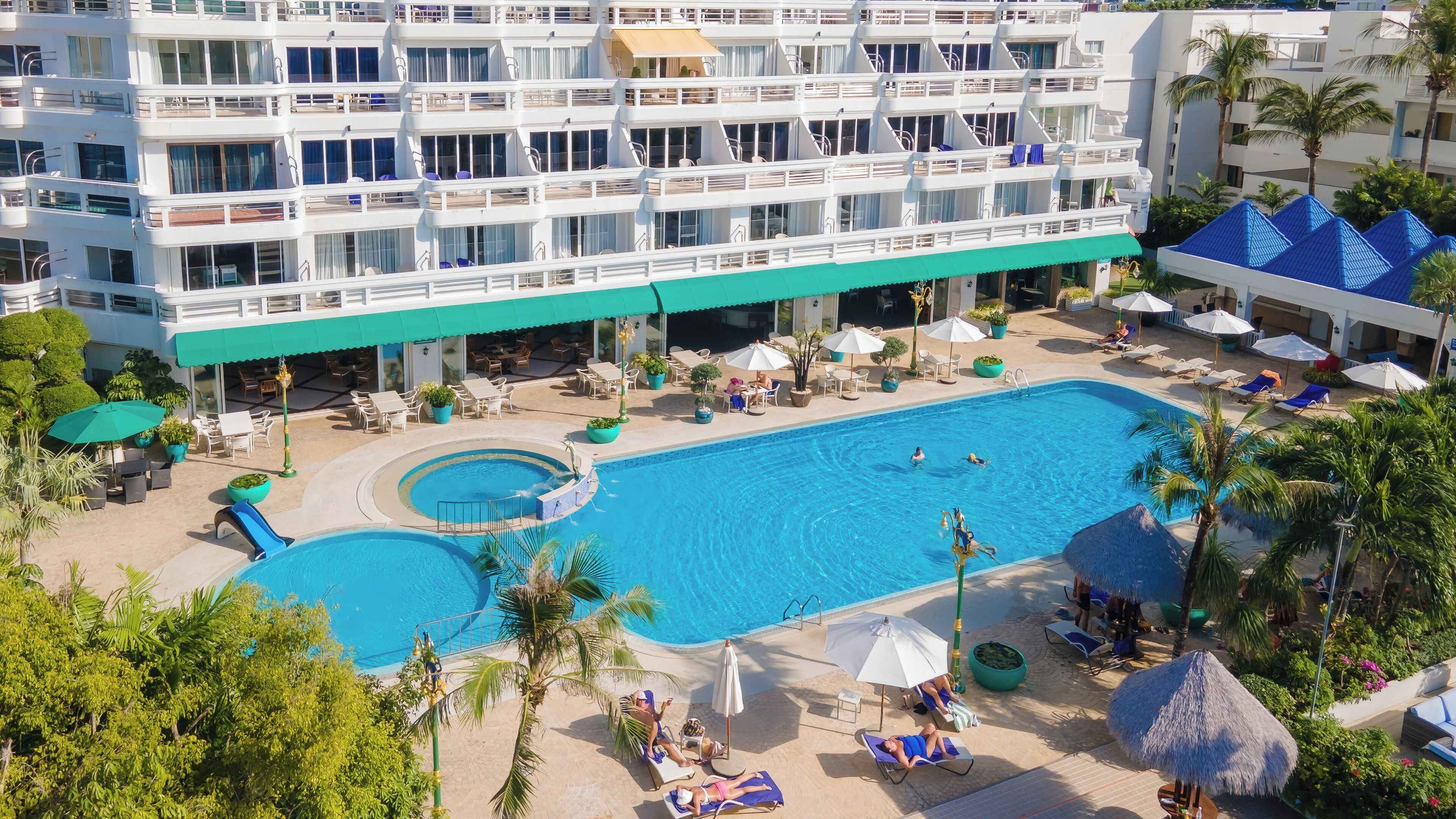 A multi-story hotel with a large swimming pool, outdoor seating, and palm trees.