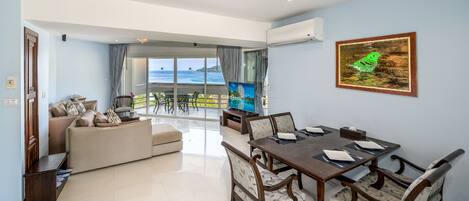 A spacious living area with a balcony overlooking the sea, a dining table set for four, and a television on a stand.