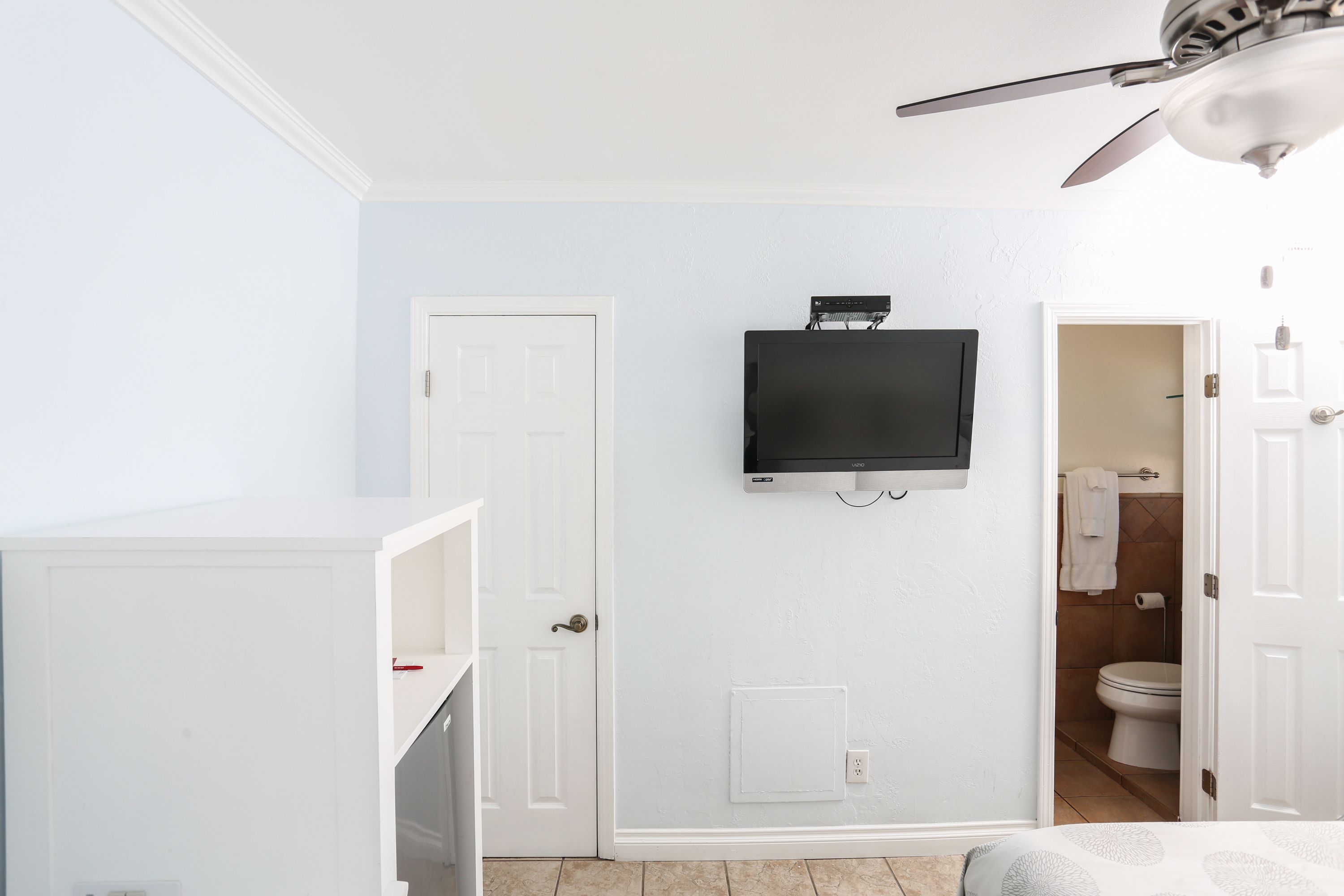 A bedroom with a TV mounted on the wall, a ceiling fan, and a door leading to a bathroom.