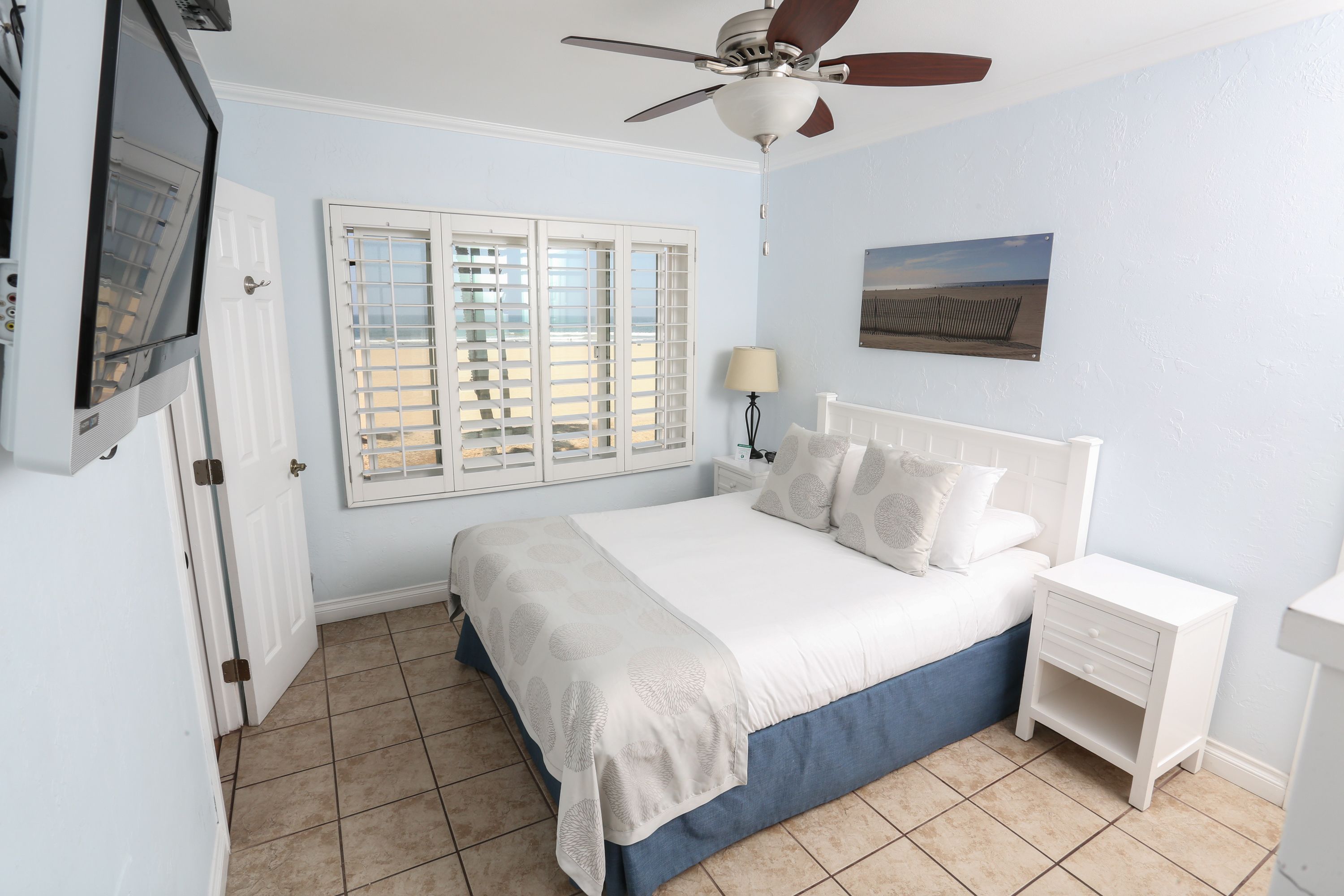 A bedroom with a bed, a nightstand, a ceiling fan, a window with shutters, and a wall-mounted picture.