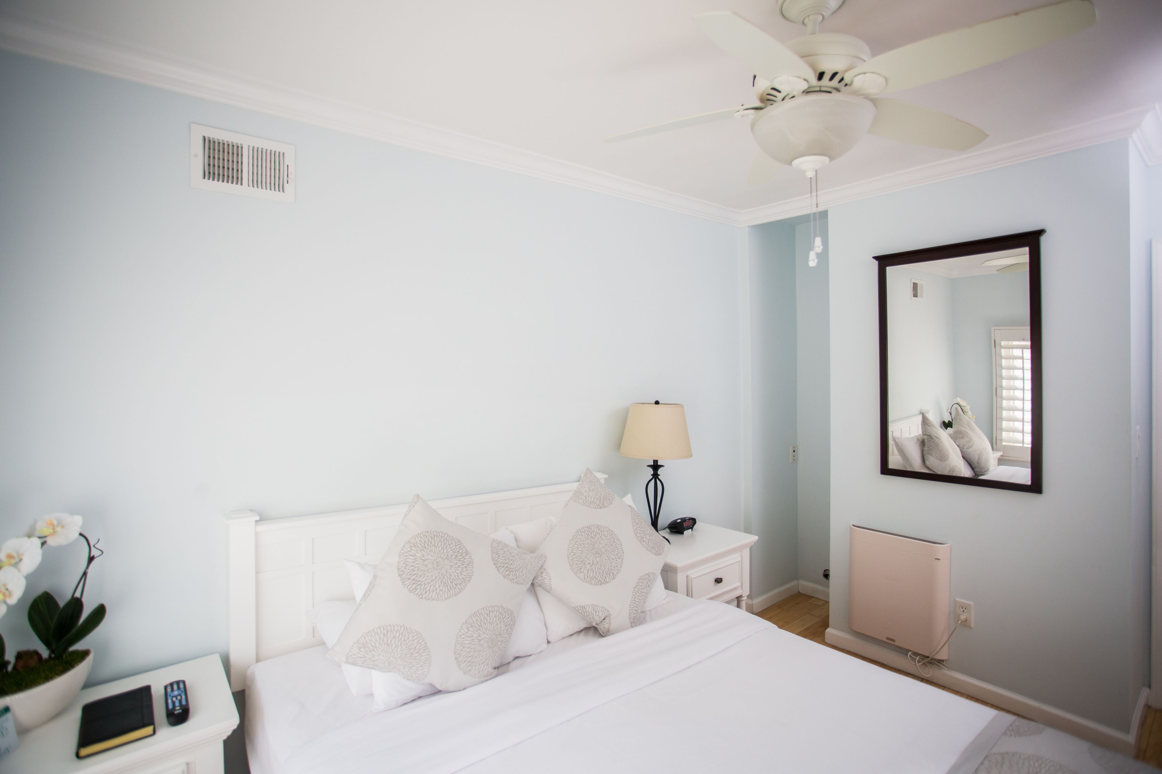 Une chambre à coucher comprenant un lit, une table de chevet, une lampe, un miroir, un ventilateur de plafond et un radiateur.