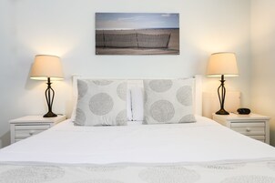 Una cama bien hecha con una colcha blanca y dos almohadas con estampado de hojas, flanqueada por dos mesitas de noche con lámparas, un cuadro en la pared con una escena de playa y una mesita de noche blanca con un cajón.