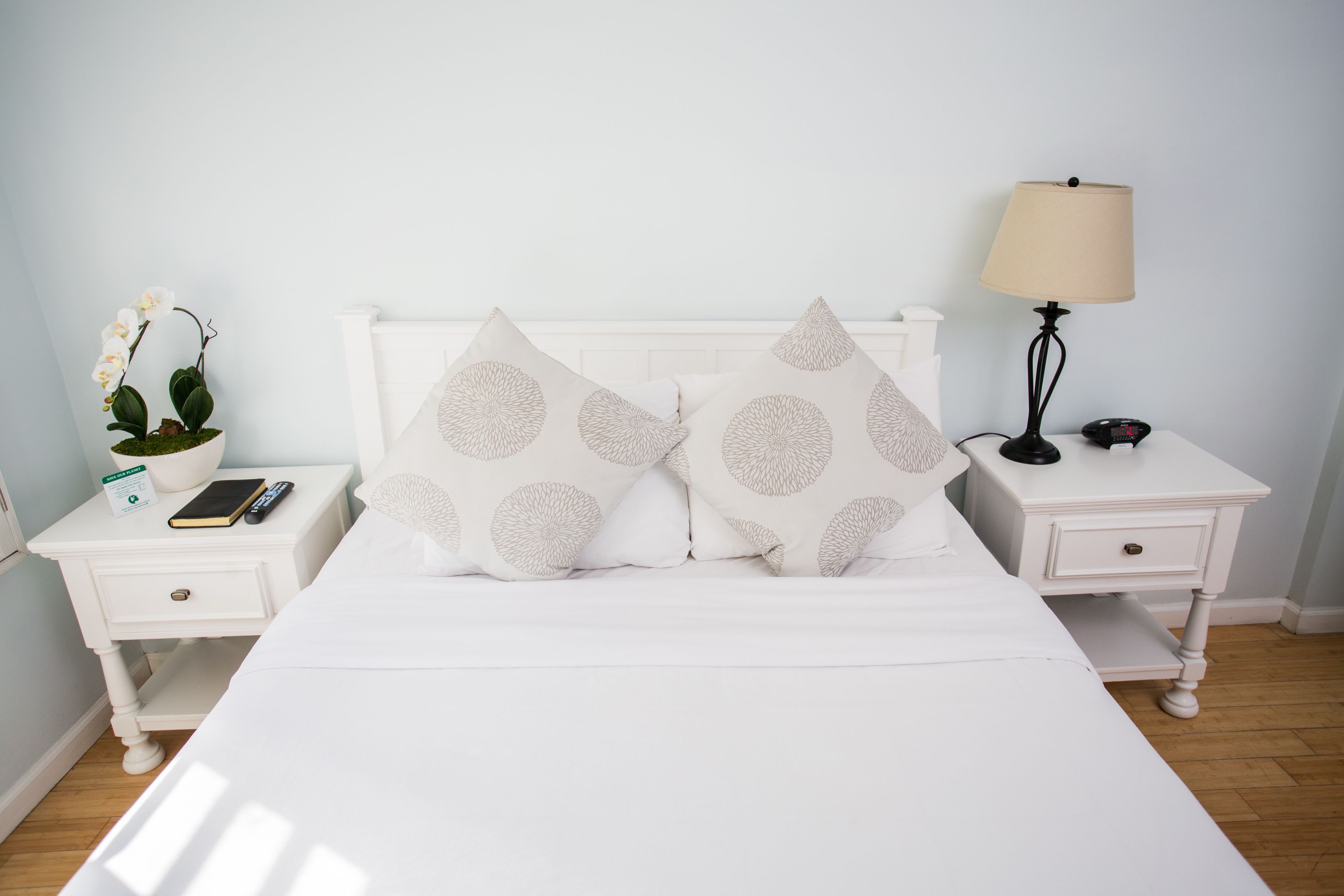 Una cama bien hecha con dos almohadas decorativas, una mesita de noche con una lámpara, un control remoto y una planta en maceta.