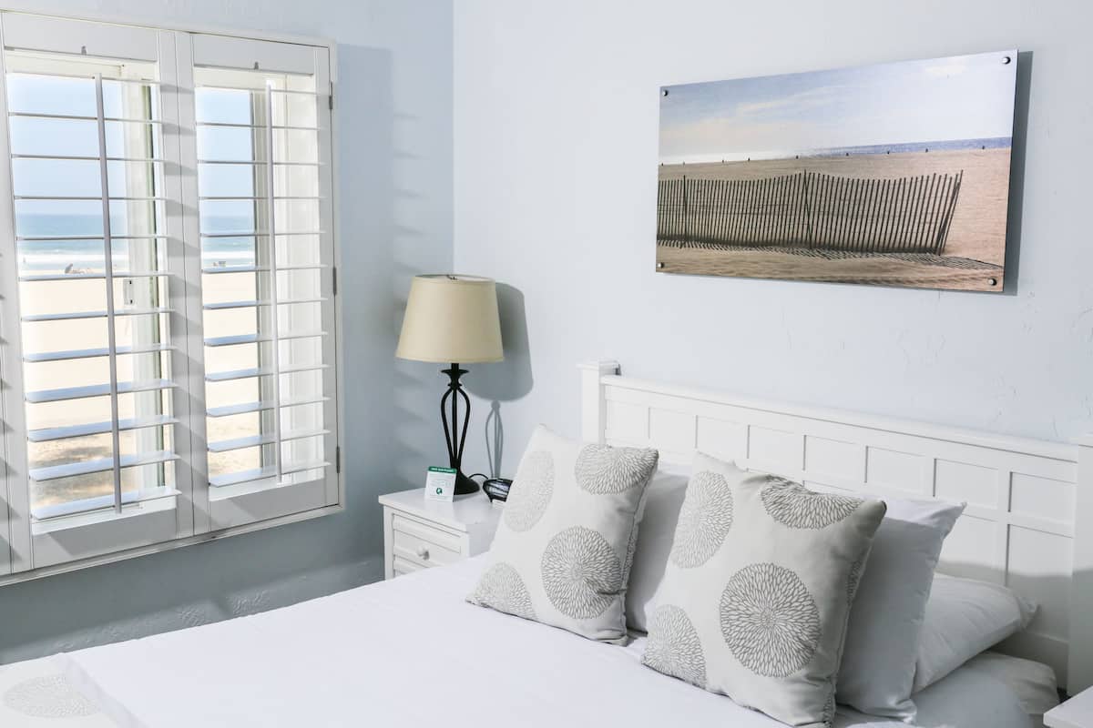 Ein Schlafzimmer mit Bett, Nachttisch in Weiß, Lampe und Blick auf den Strand durch das Fenster.