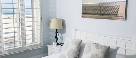 A bedroom with a bed, white nightstand, lamp, and a beach view through the window.