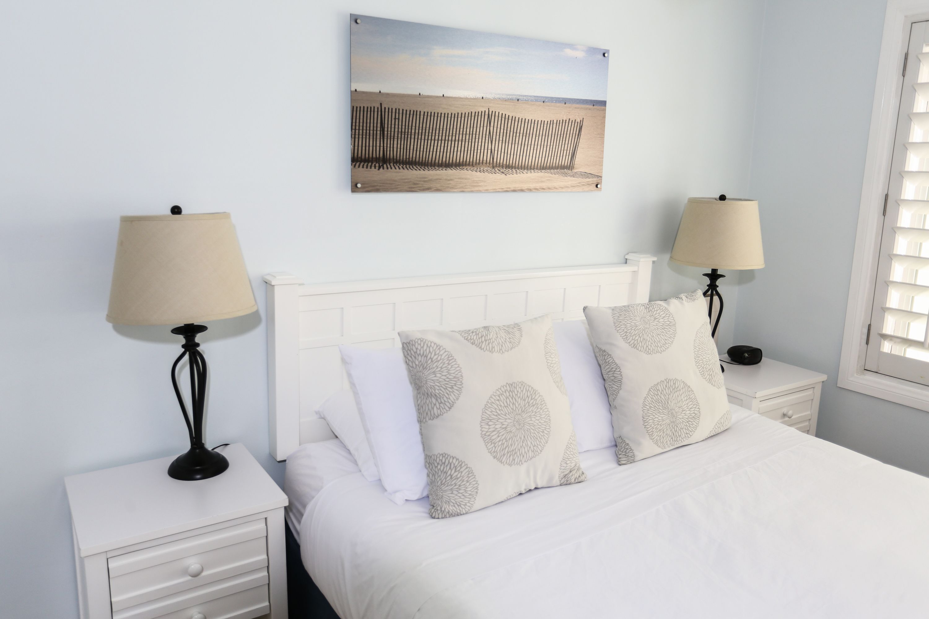 Camera da letto con un letto bianco, due comodini con lampade, un quadro appeso al muro e una finestra con persiane.