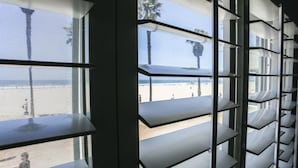 Blick durch ein Fenster mit Rollläden, der einen Strand und Palmen im Freien zeigt.