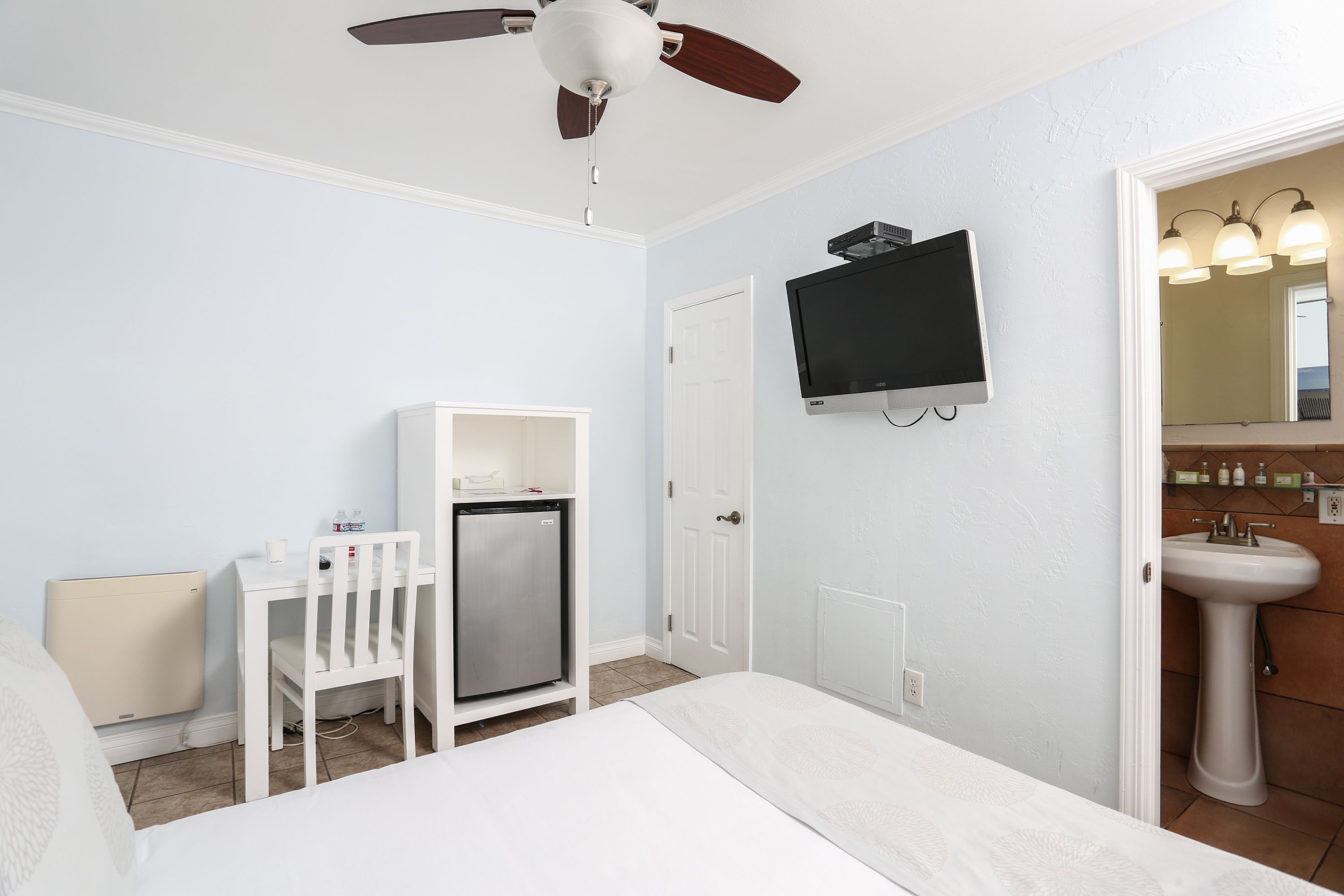 Ein Schlafzimmer mit Bett, Schreibtisch mit Stuhl, Deckenventilator, Fernseher und einem Badezimmer mit Waschbecken und Spiegel.