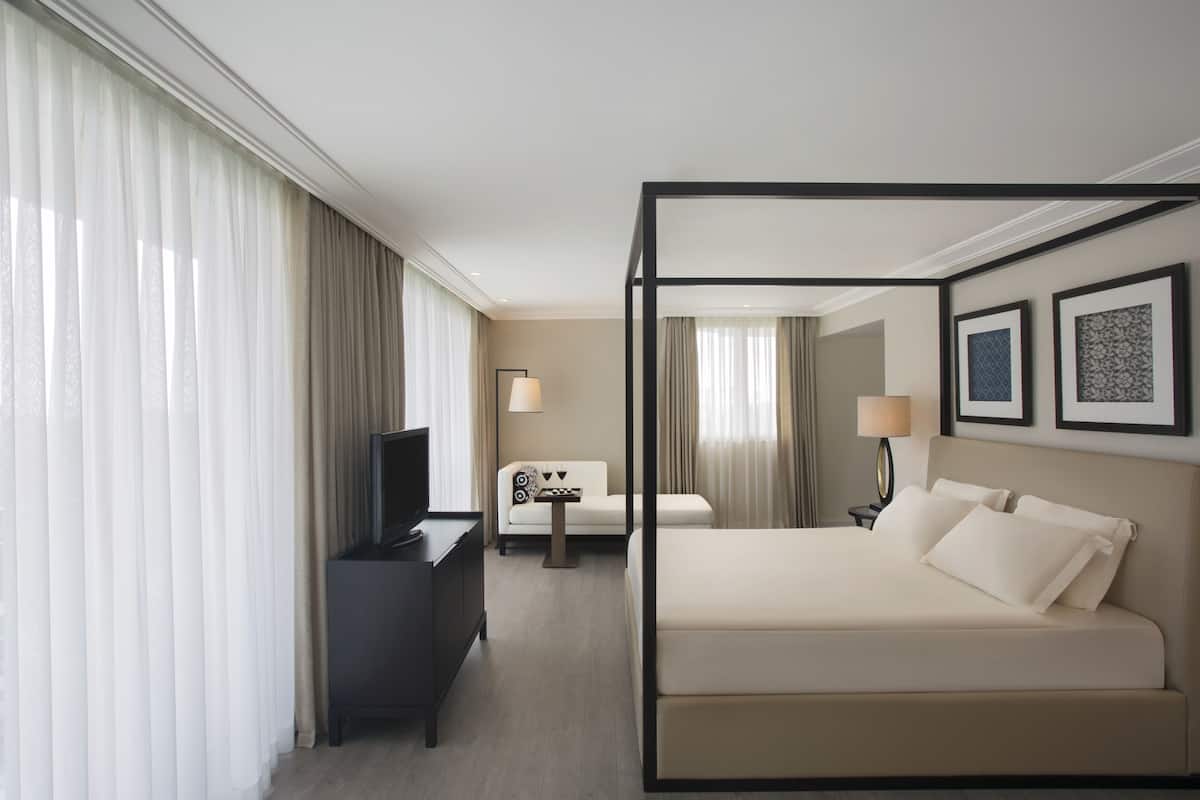 Habitación de hotel con cama con dosel, un escritorio con televisor, una silla, una mesita de noche con lámpara y dos cuadros en la pared.