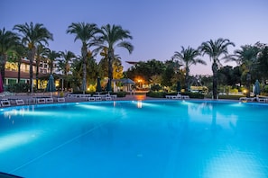 Ein Swimmingpool mit Liegestühlen und Palmen, umgeben von Gebäuden mit warmer Beleuchtung.