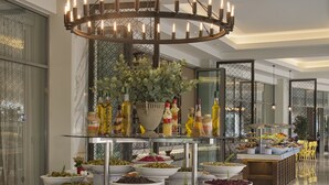 Un moderno vestíbulo de hotel con una zona de bar circular central que ofrece diversas opciones de comida y bebida, una gran lámpara de araña y una vitrina con tapa de vidrio.