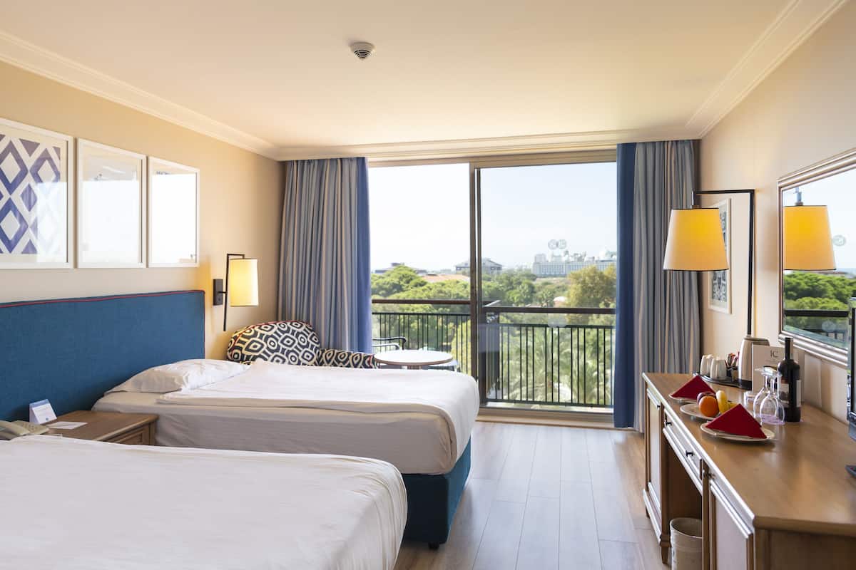 Habitación de hotel con dos camas, un balcón con vistas, una mesita con una silla y un minibar.