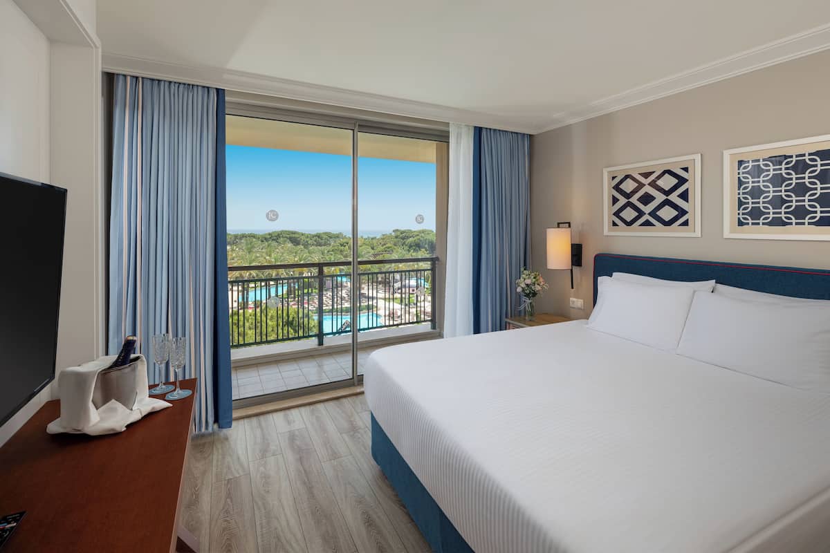 Habitación de hotel con una cama grande, televisión, escritorio con una botella y vasos, mesita de noche con lámpara, y balcón con vista a la piscina.