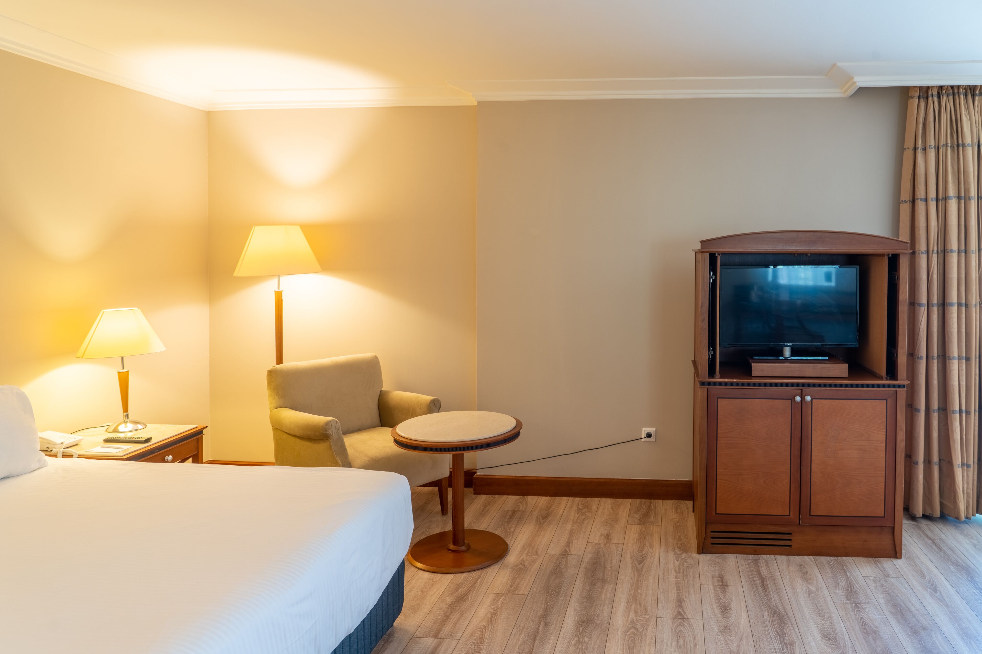A hotel room with a bed, a chair, a small table, a TV, and a lamp.