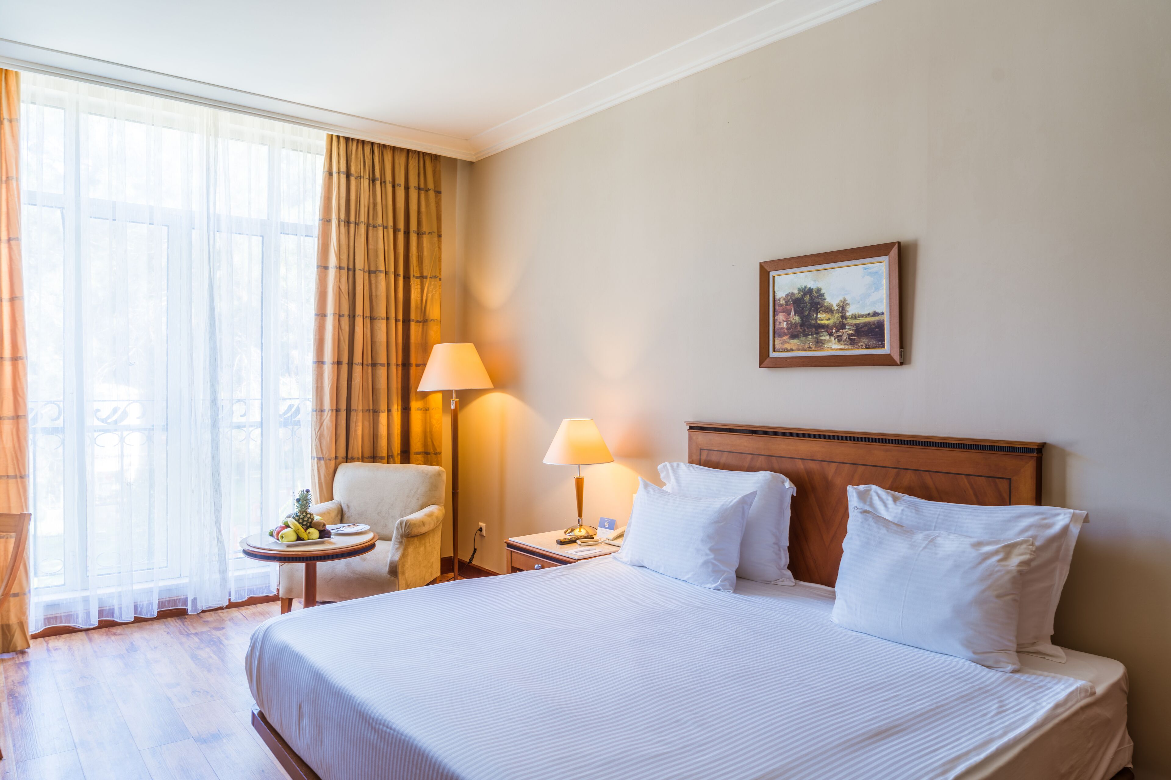 A hotel room with a large bed, a chair, a small table with fruit, a lamp, and a painting on the wall.