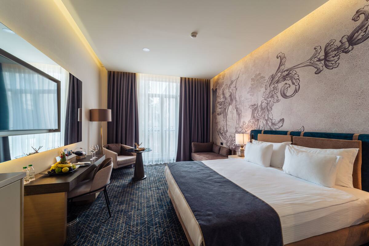 A hotel room with a large bed, a desk with a chair, a small table with a lamp, and a large window with curtains.
