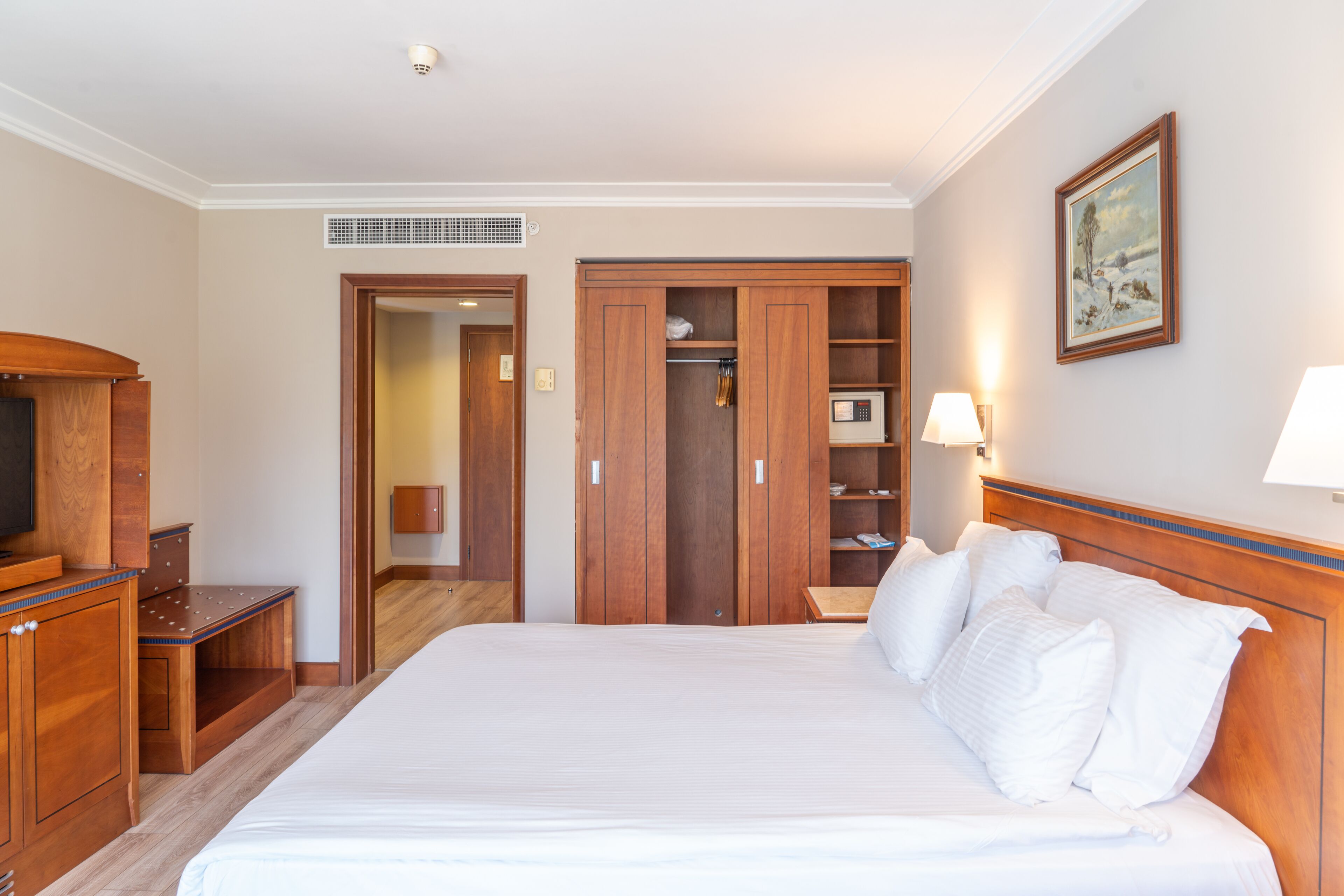 A hotel room with a large bed, wooden wardrobe, a desk with a chair, a television, and a painting on the wall.