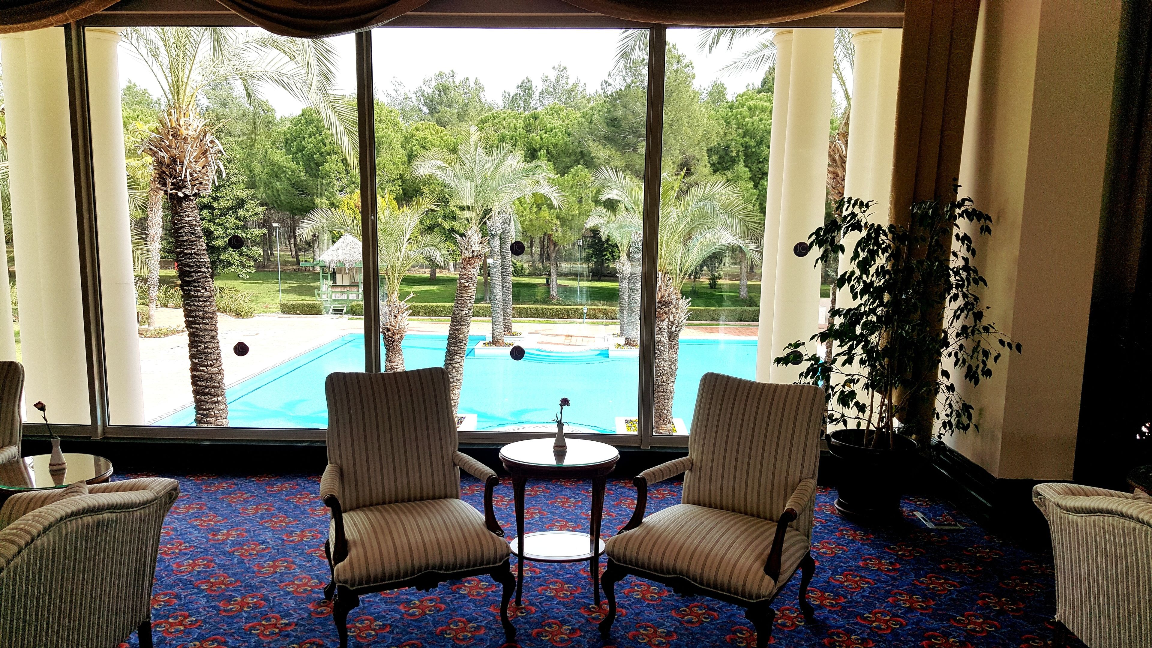 A room with a large window overlooking a pool and garden. There are two striped chairs and a small table with a vase.