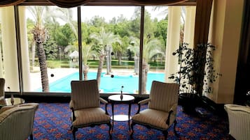 A room with a large window overlooking a pool and garden. There are two striped chairs and a small table with a vase.