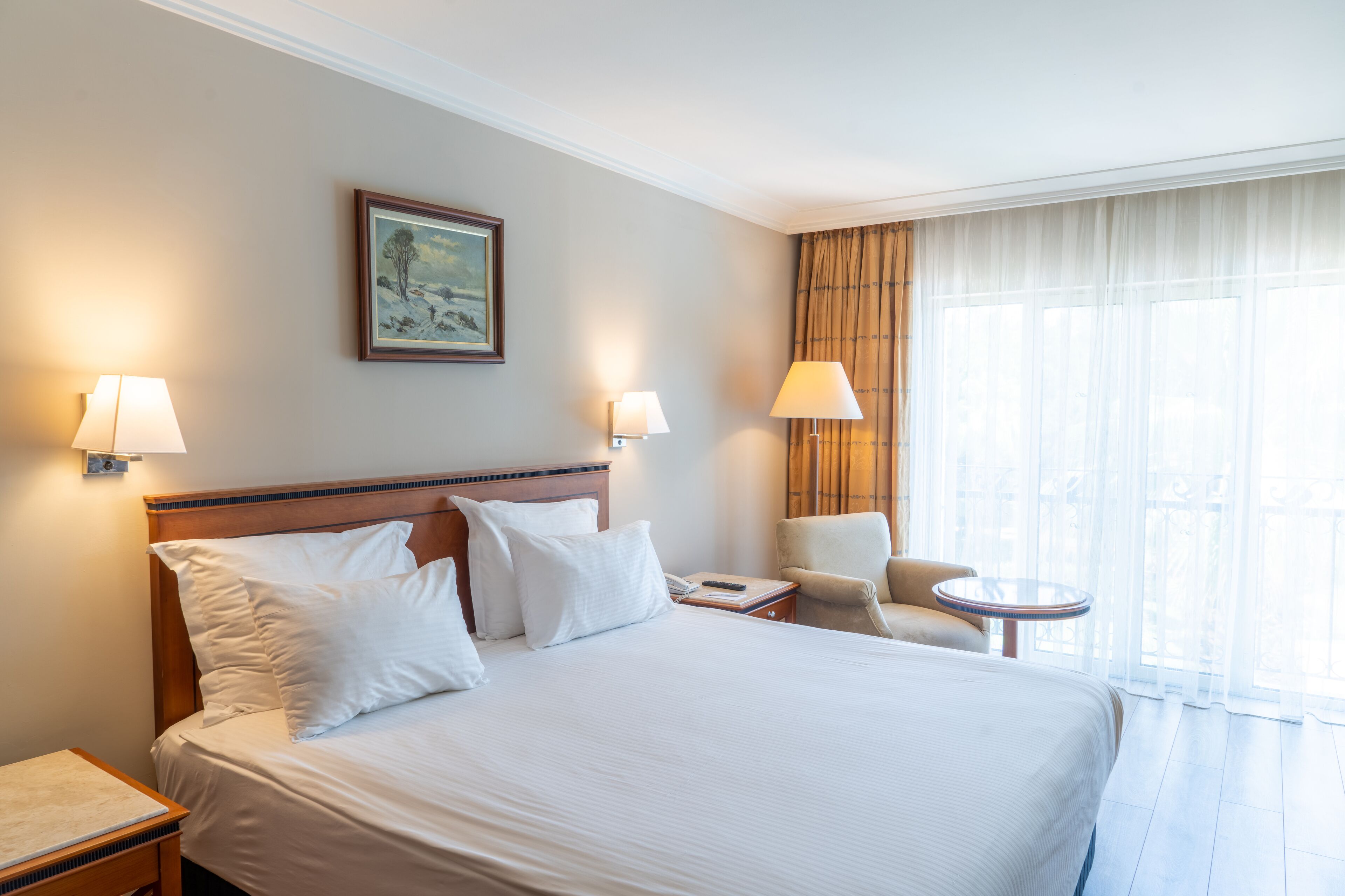 Una habitación de hotel con una cama grande, dos lámparas de noche, una silla, una mesita y un cuadro en la pared.