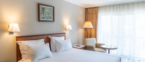 Una habitación de hotel con una cama grande, dos lámparas de noche, una silla, una mesita y un cuadro en la pared.