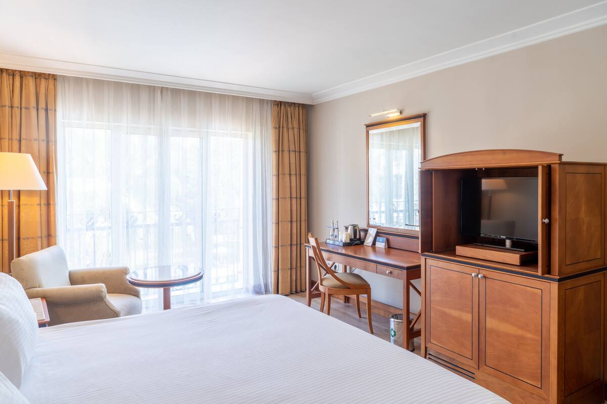A hotel room with a large bed, a desk with a television, a chair, a lamp, and a view of the outdoors through the window.