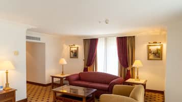 A hotel room with a sofa, armchair, and a small table. There are two lamps, a window with curtains, and a painting on the wall.