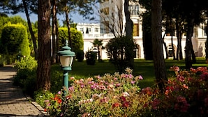 Un sentier bordé d’arbres et d’arbustes en fleurs, qui mène à un bâtiment à l’architecture classique.