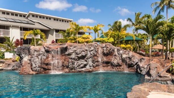 A pool with a waterfall feature surrounded by tropical plants and a building with balconies. - Oceanfront Realty - The Cliffs at Princeville (Princeville)