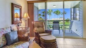 A room with a wicker chair, a sofa with a floral pattern, a lamp, and a view of palm trees through a sliding glass door. - Oceanfront Realty - The Cliffs at Princeville (Princeville)