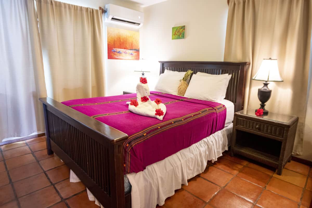 Une chambre d’hôtel avec un lit en bois, une couvre-lit violette, des oreillers blancs, une table de chevet avec une lampe et un tableau accroché au mur.