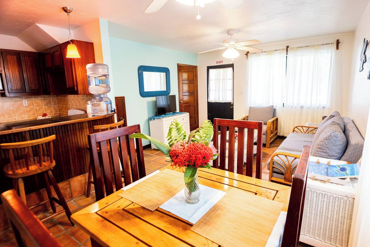Un coin repas avec une table en bois, des chaises, un vase de fleurs et, au fond, une cuisine.