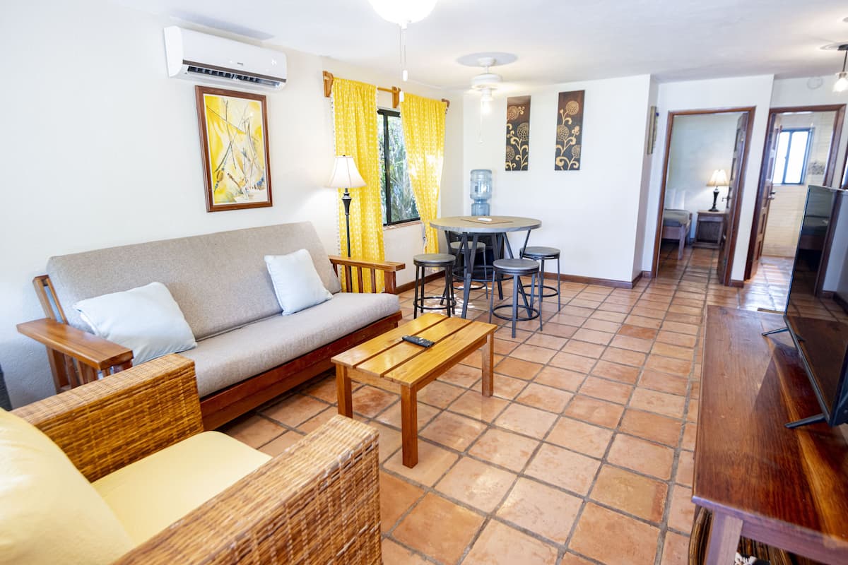 Un salon comprenant un canapé, une table basse et des meubles en osier. On y trouve également un coin repas avec une table et des chaises, ainsi qu’un distributeur d’eau.