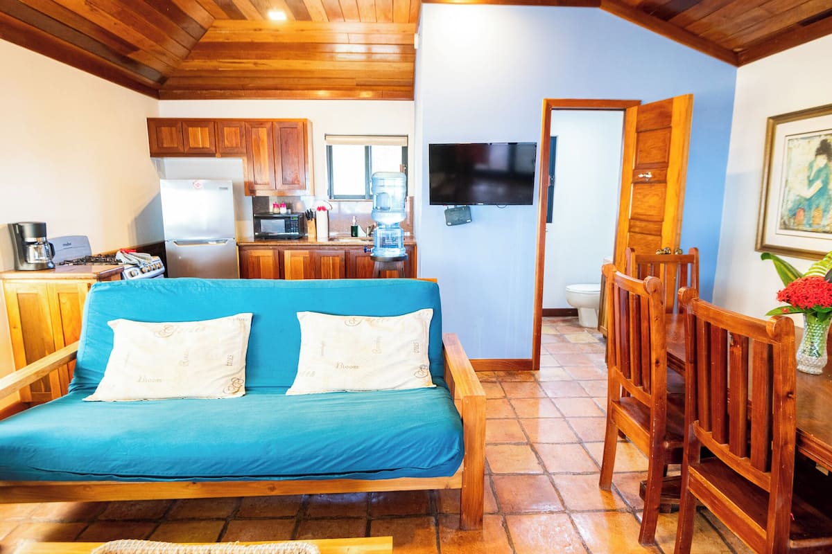 Un salon chaleureux avec un canapé bleu, des meubles en bois, une cuisine dotée de placards en bois, un téléviseur fixé au mur et un sol carrelé.