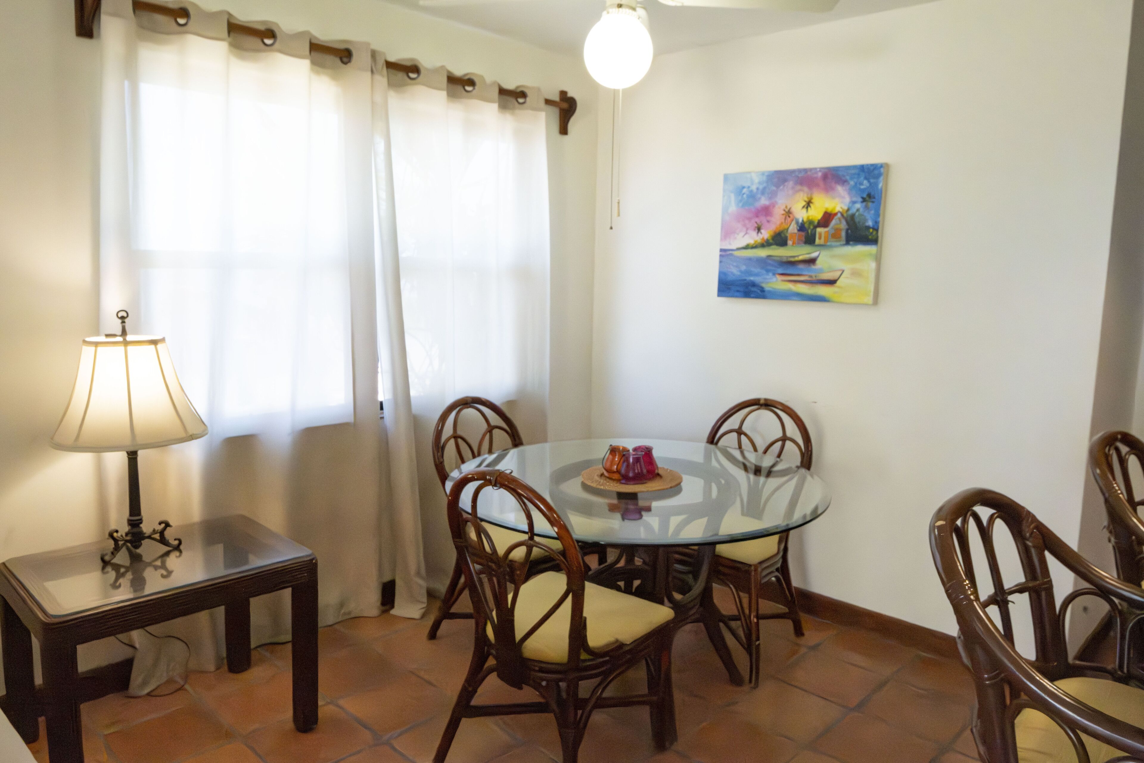 A dining area with a round glass table, four wicker chairs, a small side table with a lamp, and a painting on the wall.