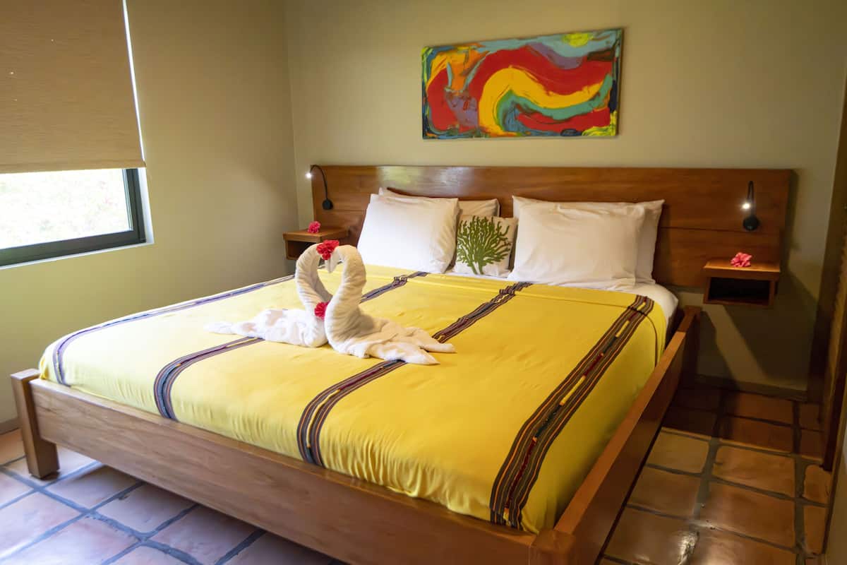 Un lit bien fait, avec une couvre-lit jaune et des cygnes décoratifs, une tête de lit en bois et une peinture abstraite aux couleurs vives accrochée au mur.
