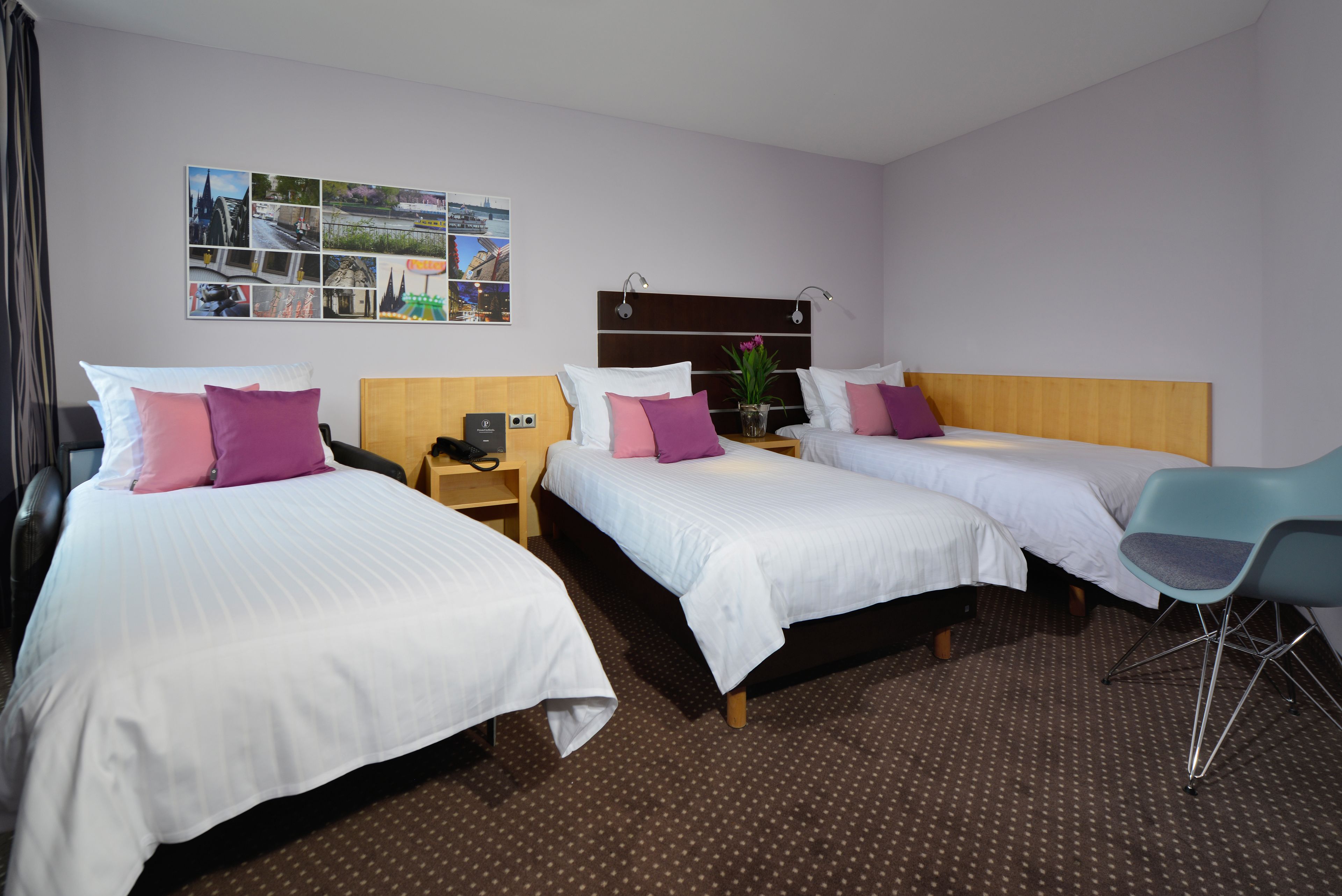 Une chambre d’hôtel avec trois lits simples, une tête de lit en bois, une table de chevet avec une lampe, une chaise et un collage de photos accroché au mur.