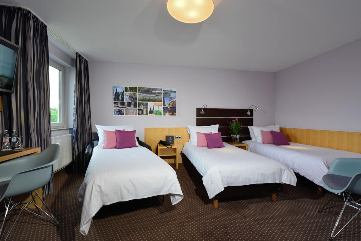 A hotel room with two single beds, a desk with a chair, a small table with a lamp, and a wall-mounted TV.