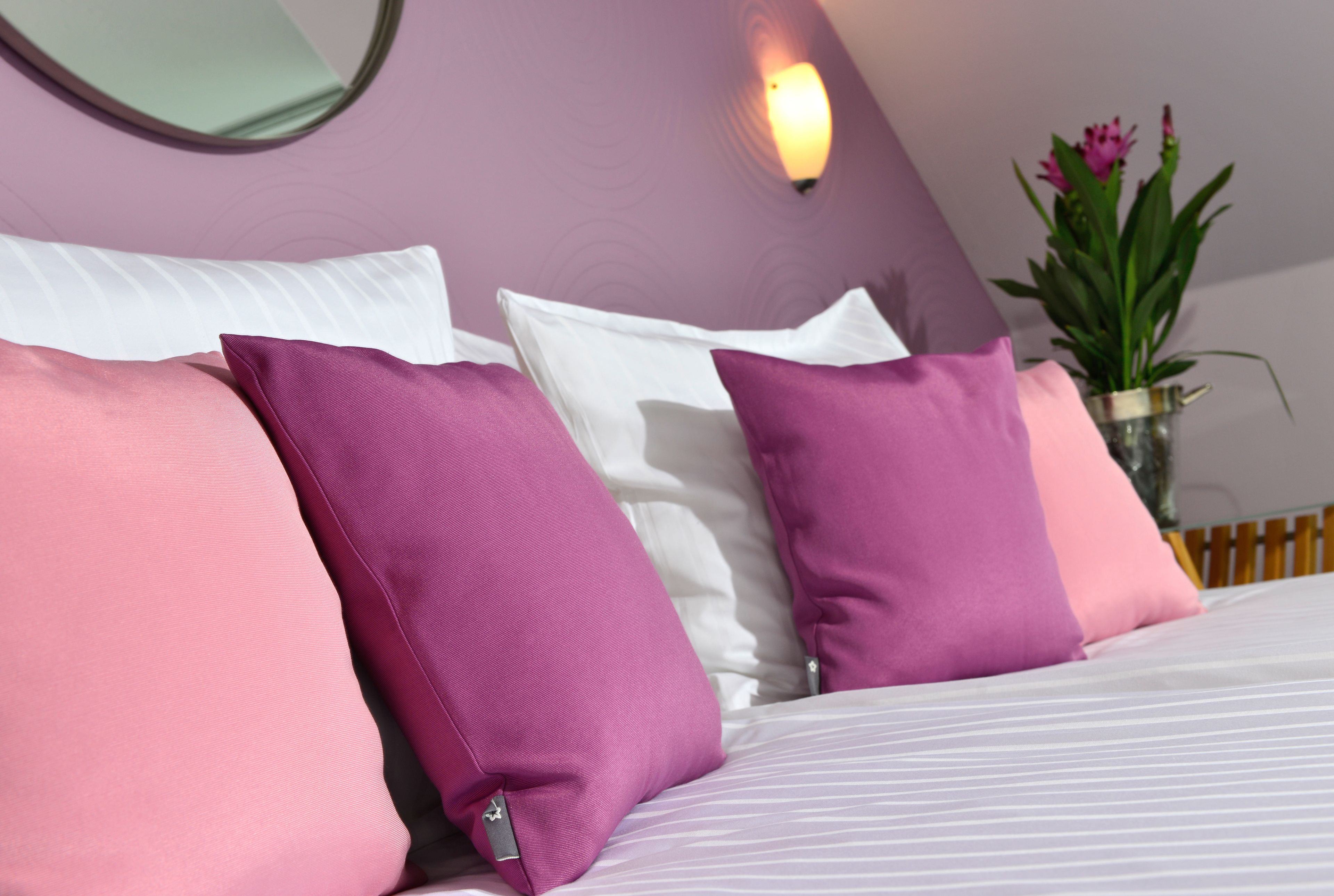 Una cama con almohadas blancas y rosadas, un florero con flores y una lámpara fijada a la pared.