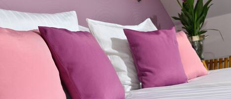 Una cama con almohadas blancas y rosadas, un florero con flores y una lámpara fijada a la pared.