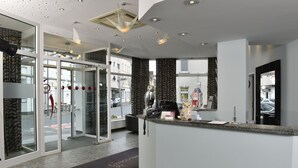 A hotel reception area with a glass door entrance, a reception desk, and a view of the street outside. - Hotel UHU Garni Köln (Cologne)