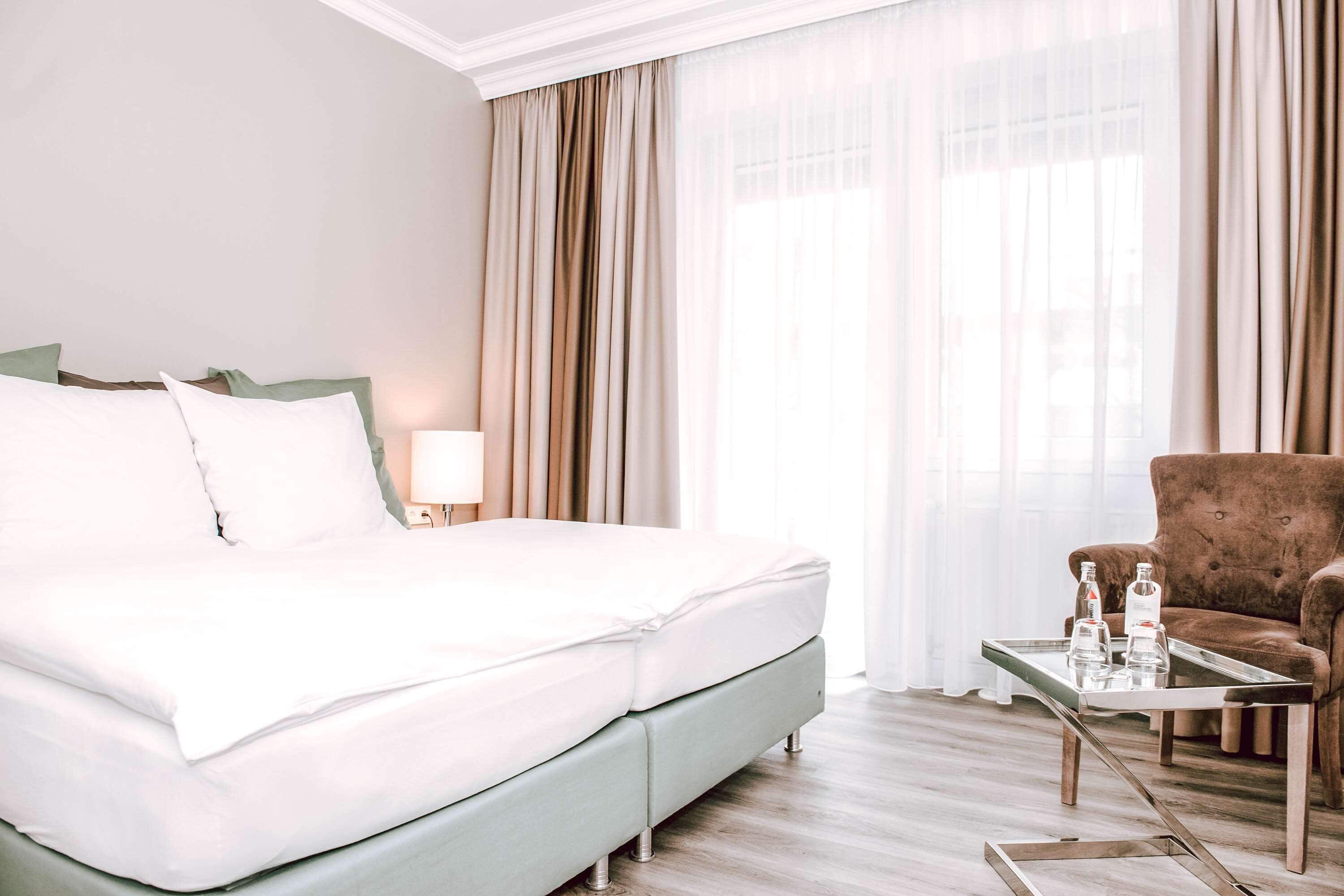 A hotel room with a bed, a chair, a table with bottles, and a window with curtains.
