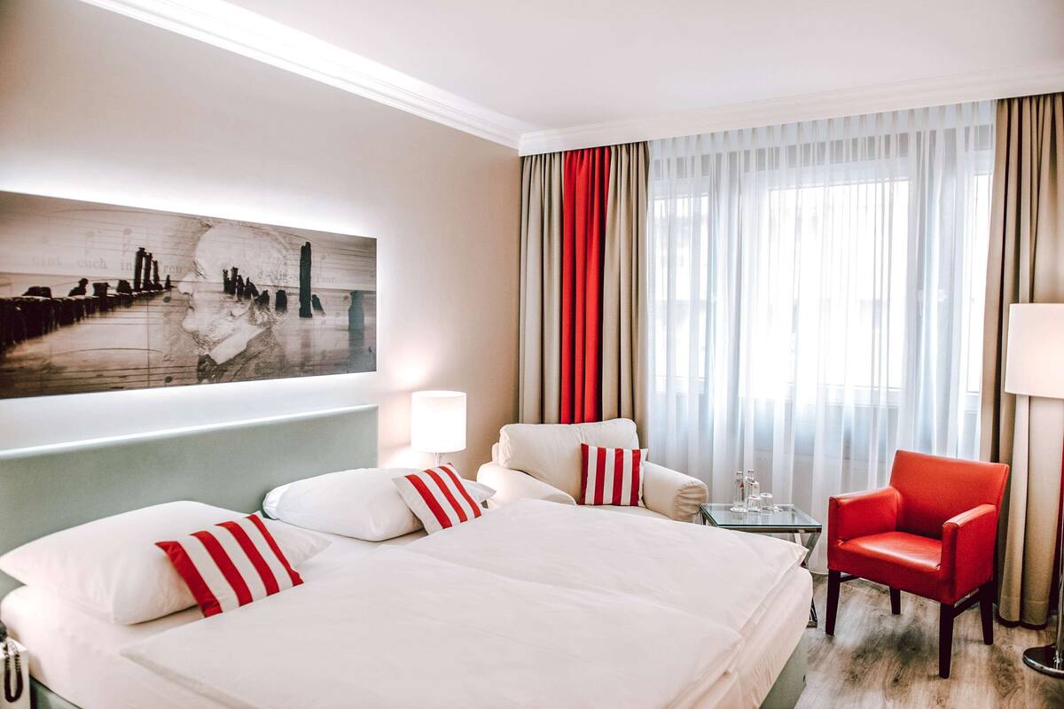 A hotel room with a large bed, two armchairs, a red chair, a glass table, and a painting on the wall.